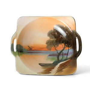 May include: Square ceramic serving tray with a painted landscape scene. The tray features a sunset over a body of water with trees and a small boat. The tray has two handles and a cream-colored border.