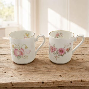 May include: Two white ceramic mugs featuring delicate floral patterns. The left mug displays pink and yellow sweet pea flowers, while the right mug showcases a cluster of pink dahlias and small blossoms. Both mugs have gold-trimmed rims and handles.