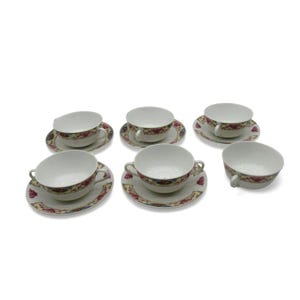 May include: A set of six white teacups and saucers. Each cup has a handle and is decorated with a floral pattern in red, yellow, and blue. The saucers match the cups' design.