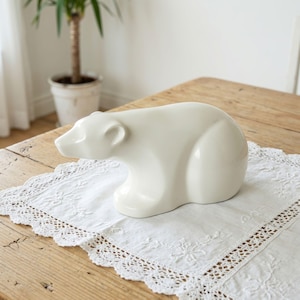 Könnte beinhalten: Eine glatte, cremeweiße Keramikfigur eines Eisbären in Laufpose. Die Skulptur hat eine glänzende Oberfläche, einen abgerundeten Rücken und eine ausgeprägte Schnauze mit kleinen Ohren, die das Tier in einer vereinfachten, massiven Form darstellt.