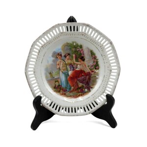 May include: White, octagonal decorative plate with a pierced border. The center features a painted scene of three women in classical attire, surrounded by flowers and a column. The plate is displayed on a black stand.