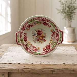 May include: A round ceramic bowl with two side handles, featuring a cream-colored base decorated with a vibrant floral pattern in shades of deep red, pink, yellow, and green. The rim has a textured, rope-like edge and a detailed botanical border.