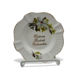 May include: White ceramic ashtray with a ruffled edge and gold trim. The ashtray features a floral design with green and yellow accents and the text "Victoria British Columbia". The ashtray is on a white metal stand.