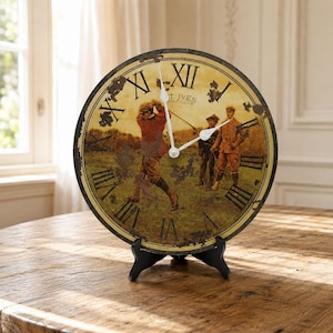 Op de afbeelding: Een ronde wandklok met een vintage-stijl illustratie van golfers op een baan met de tekst St. Ives Golf Club. De wijzerplaat heeft zwarte Romeinse cijfers, een verweerde afwerking en witte wijzers, geplaatst op een zwarte kunststof standaard.