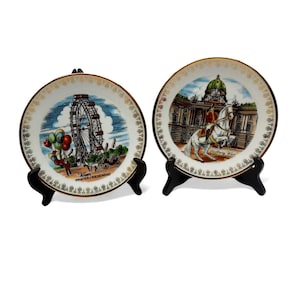 May include: Two decorative ceramic plates with gold rims and black stands. One plate features a Ferris wheel with balloons, the text "Wien Prater/Riesenrad". The other plate depicts a building and a person on a horse. Both plates have a decorative border.