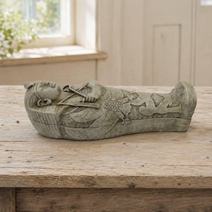 May include: A light gray stone-colored sarcophagus figurine featuring an Egyptian-style figure with arms crossed over the chest. The surface is decorated with intricate carved wing patterns and linear details, presenting a weathered, textured finish.