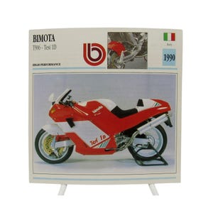 Pode incluir: Uma placa retangular com uma motocicleta Bimota T906 Tesi 1D vermelha e branca. O texto diz Bimota T906 - Tesi 1D, High Performance, 1990 e Italy. Uma pequena imagem inserida mostra a roda traseira e a suspensão da motocicleta.