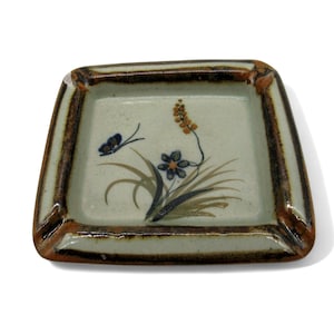 May include: A ceramic ashtray with a brown rim and a white interior. The ashtray features a hand-painted design of flowers and butterflies in shades of blue, green, and brown.