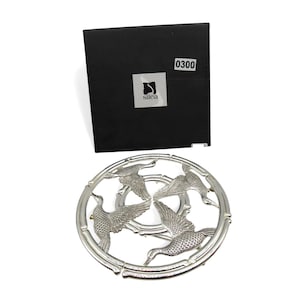 May include: Silver metal trivet with a design of four flying ducks within a circular bamboo-style frame. A black box with the number "0300" and the "silea" logo is in the background. Suitable for kitchen or dining use.
