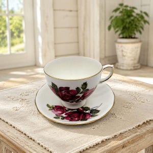 May include: A white porcelain teacup and saucer set featuring a detailed floral pattern of deep red roses with green leaves. The teacup has a curved handle and both the cup and saucer are finished with a thin gold trim along the edges.