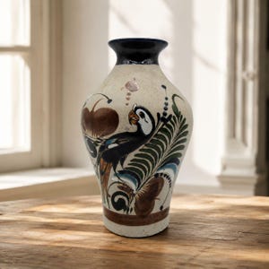 May include: A ceramic vase with a cream-colored base and a dark blue rim. The exterior features hand-painted illustrations of a bird with black, white, and orange plumage, surrounded by green fern-like leaves and brown circular floral motifs.