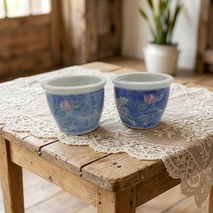 May include: Two small ceramic cups with a white interior and a blue exterior featuring a hand-painted floral pattern of pink roses and green leaves. The cups have a rounded shape with a smooth rim and a tapered base, showcasing a glossy finish.