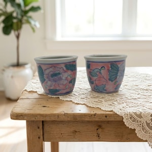 Könnte beinhalten: Zwei kleine Keramikbecher mit einem handgemalten Blumenmuster in zartem Rosa, Türkis und Weiß. Die Becher haben eine abgerundete Form mit einem glatten Rand und einem sich verjüngenden Boden und zeigen detaillierte botanische Blütenmotive.