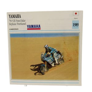 May include: A vintage Yamaha 750 YZE Paris-Dakar racing card featuring Stéphane Peterhansel. The image shows a blue and white motorcycle racing through a desert landscape. The card includes the year 1989 and the Japanese flag.
