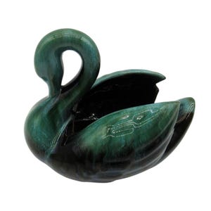 May include: A green and black glazed ceramic swan figurine. The swan has a long neck and a wide wingspan. The glaze is a mottled green and black, with a hint of blue.