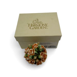May include: A decorative floral sculpture featuring a cluster of orange roses with green leaves and buds. The sculpture is presented with a beige box that has the text "Lord Byron's Harmony Garden" printed on it.