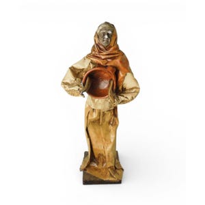 May include: A handcrafted figurine of a woman wearing a headscarf and long dress, holding a brown bowl. The figure is made of paper and has a weathered appearance, standing on a wooden base. The color palette includes shades of brown and tan.