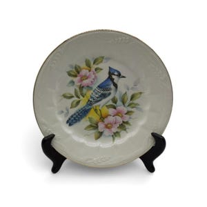 May include: A decorative white plate featuring a blue jay perched among pink flowers and green leaves. The plate has a gold rim and is displayed on a black stand. The plate's design is a painted illustration.