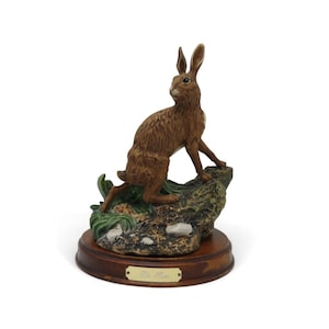 Peut inclure: Une figurine de lièvre brun debout sur une base rocheuse texturée avec du feuillage vert et des pierres blanches. La sculpture est montée sur un socle circulaire en bois avec une petite plaque métallique dorée portant le texte The Hare.