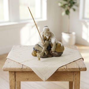 May include: A porcelain figurine of an elderly person with a white beard, wearing a white shirt and blue pants, sitting on a rock while holding a long wooden fishing rod with a small fish attached to the line and a small brown jug nearby.