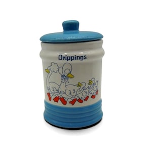 May include: A vintage ceramic jar with a blue lid and base, and the word "Drippings" in black text. The jar features a whimsical illustration of ducks in blue, yellow, and red. A charming kitchenware item.