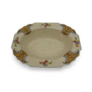 Puede incluir: Plato ovalado de cerámica color crema con borde festoneado. El plato presenta detalles decorativos amarillos y negros en las esquinas y racimos florales en tonos rojos, amarillos y morados. El plato tiene un esmalte craquelado.