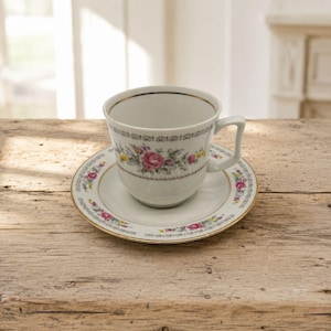 Może przedstawiać: Biała porcelanowa filiżanka do herbaty ze spodkiem, ozdobiona delikatnym kwiatowym wzorem z różowymi różami i żółtymi kwiatami, podkreślona cienką złotą obwódką oraz dekoracyjnym wzorem wokół środka filiżanki i na krawędzi spodka.