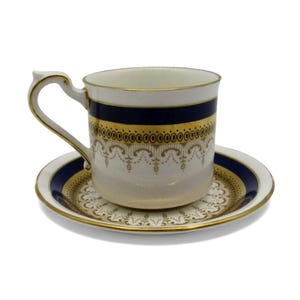 Puede incluir: Un juego de taza y platillo blanco con ribete dorado y bandas azul marino. La taza y el platillo presentan detalles dorados ornamentados y un asa delicada. El juego está diseñado para el servicio de té o café.