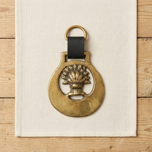 Pode incluir: Um ornamento de latão para cavalo com um design central em relevo de um feixe de trigo. A peça metálica circular está presa a uma tira de couro preta com um anel de latão no topo, criando um ornamento decorativo suspenso com acabamento metálico.