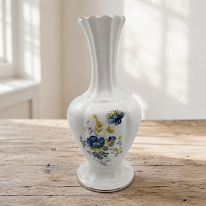 May include: A white porcelain vase with a scalloped rim and a tapered neck. The body features a floral pattern with blue blossoms, green leaves, and yellow accents. The base is circular and flared, with a thin gold trim along the bottom edge.