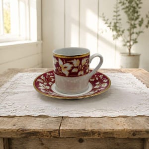 May include: A porcelain teacup and matching saucer featuring a deep burgundy band decorated with white floral patterns and gold trim. The cup has a curved handle and a smooth, glossy finish, while the saucer displays a coordinating floral motif.