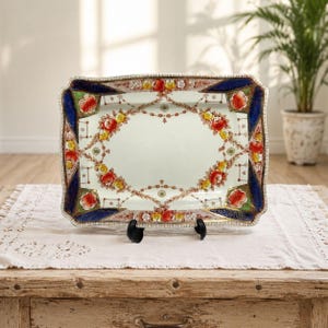 May include: A rectangular ceramic serving tray featuring a cream-coloured centre surrounded by a decorative border of red and yellow floral garlands. The corners are accented with deep blue panels and green details, finished with a gold-toned trim.