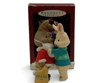 May include: A Keepsake Ornament featuring two rabbits. One rabbit wears a red coat with white trim and holds a tan sack labelled Mom & Dad 1994. The other rabbit wears a teal sweater with white star patterns. The ornament stands before its original box.