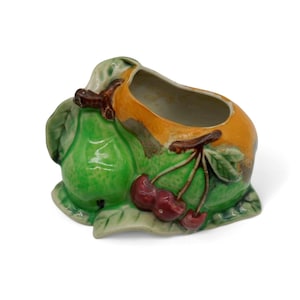 Op de afbeelding: Een keramische plantenbak in de vorm van een groep fruit, met een felgroene peer, een glad oranje gedeelte en drie kleine rode kersen met bruine steeltjes. Het object heeft een glanzende afwerking en een holle kern voor kleine voorwerpen.