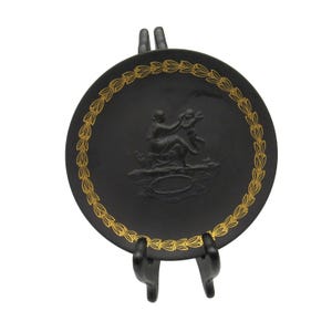 May include: A black decorative plate with a gold leaf border and a raised relief of two figures. The plate is displayed on a black stand, showcasing its intricate design and classic aesthetic.