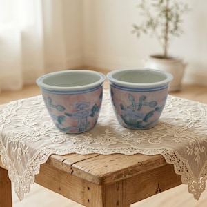 Könnte beinhalten: Zwei Keramikschalen mit einem zartrosa und weiß marmorierten Hintergrund und handgemalten blauen und grünen Blumenmotiven. Jede Schale hat einen glatten, abgerundeten Rand und einen konischen Boden, was ein feines künstlerisches Design zeigt.