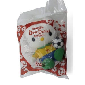 May include: A reversible Dear Daniel McFlurry plush toy in a clear plastic bag. The plush is white with a yellow and blue outfit, green shoes, and a soccer ball. The bag has red and white soccer ball graphics and the word "Brazil".