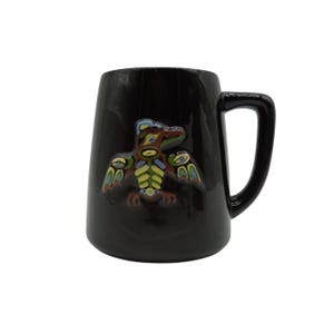 Peut inclure: Mug en céramique noire avec une anse, orné d'un motif en relief coloré d'un oiseau stylisé. L'oiseau présente des accents bleus, jaunes et verts. La tasse a une finition brillante et une anse simple et incurvée.