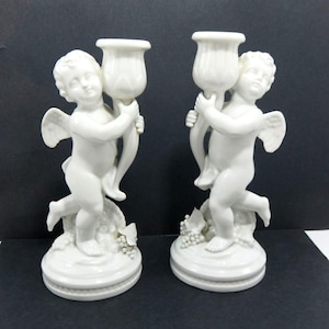 May include: A pair of white ceramic cherub candle holders. Each cherub holds a tulip-shaped candle cup. The cherubs stand on round bases with grape and leaf details. These candle holders are a classic decorative item.