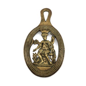 Puede incluir: Una placa de latón de forma ovalada con un diseño en relieve de una figura con túnica que sostiene un bastón con serpientes entrelazadas. La placa tiene un acabado metálico envejecido y texturizado con un pequeño lazo para colgar.