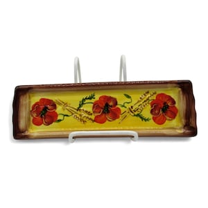 Könnte beinhalten: Rechteckige Keramikschale mit gelber Mitte, braunem Rand und rotem Mohnblumen-Design. Die Schale wird von einem weißen Metallständer getragen. Die Mohnblumen sind mit grünen Blättern und Weizenähren bemalt.