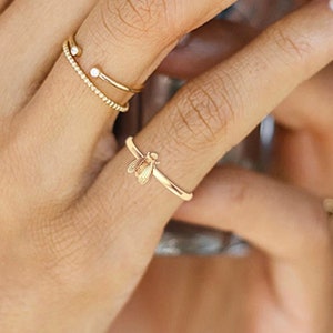 May include: A close-up of a hand wearing three gold rings. The ring on the index finger has two small diamonds. The ring on the middle finger is a thin band. The ring on the ring finger is a delicate bee design.