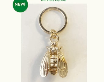 Può includere: Portachiavi a forma di ape color oro con anello portachiavi rotondo. Il portachiavi riporta la scritta "BOW TO THE BEE" e "BEE KIND, Keychain". Un cerchio verde con "NEW!" è in alto a sinistra.