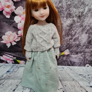 May include: A doll with long red hair wearing a grey sweater and a green skirt with a dandelion print.