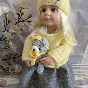 Könnte beinhalten: Puppe mit gelber Strickmütze und Pullover, grauem Rock, hält einen gehäkelten Teddybären in Grau und Gelb. Die Puppe hat lange blonde Haare und weiße Schuhe. Blumiger Hintergrund.