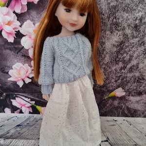 Può includere: Bambola con lunghi capelli rossi, indossa un maglione a trecce azzurro chiaro e una gonna bianca a pois argentati. La bambola ha un incarnato chiaro ed è posta davanti a uno sfondo floreale.