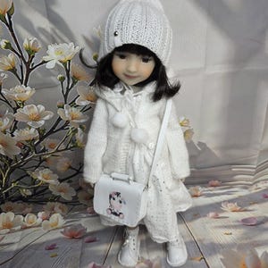 Puede incluir: Una muñeca vestida con un gorro, abrigo y vestido de punto blanco, con un pequeño bolso blanco con una vaca de dibujos animados. La muñeca lleva botas blancas y posa sobre una superficie de madera con pétalos de flores esparcidos.