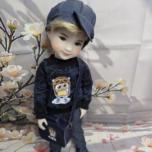 Könnte beinhalten: Puppe in einem marineblauen Outfit: Mütze, Pullover mit Eulenmotiv, Hose, Schuhe und Tasche. Die Puppe steht vor einem floralen Hintergrund.