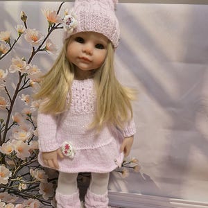 Peut inclure: Poupée vêtue d'une tenue tricotée rose clair, comprenant un bonnet à pompon, un pull, des jambières et des chaussures. La poupée a de longs cheveux blonds et porte des collants blancs. La tenue est ornée de motifs floraux.