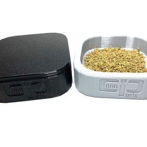 Könnte beinhalten: Ein schwarzer und ein grauer quadratischer Behälter mit dem Text "cana arts" auf der Unterseite. Der graue Behälter enthält eine kleine Menge getrockneter Kräuter.